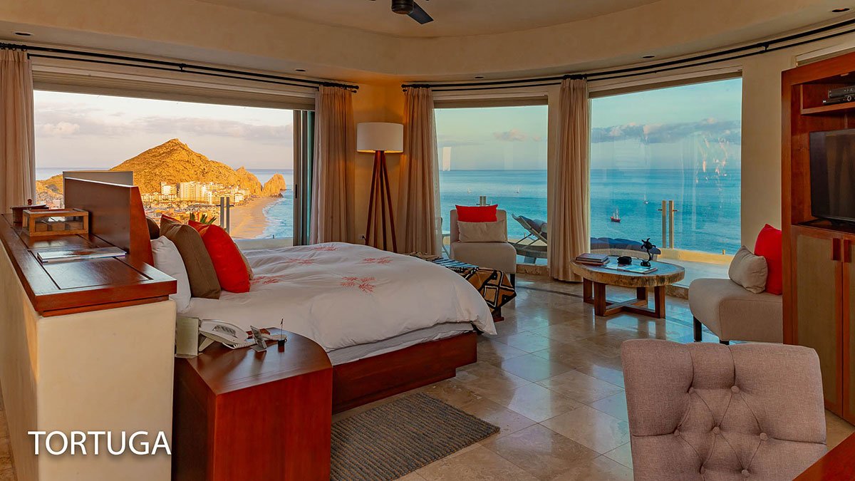 Luxury hotel room with panoramic ocean view, featuring a large bed, modern furniture, and two floor-to-ceiling windows showcasing a beach and mountain landscape at sunset.