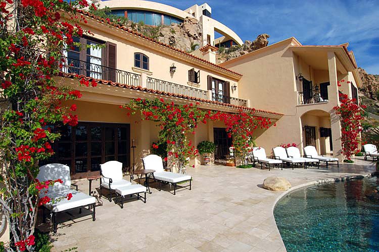 Luxury villa with poolside lounge chairs, flowering vines on walls, and a stone patio, set against a mountainous backdrop.