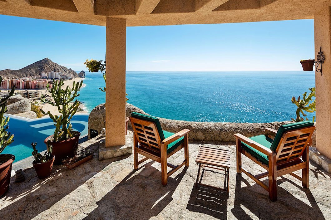 Scenic vista from a patio with two wooden chairs, a small table, potted cacti, and a view of a beach, ocean, and distant mountains under a blue sky.
