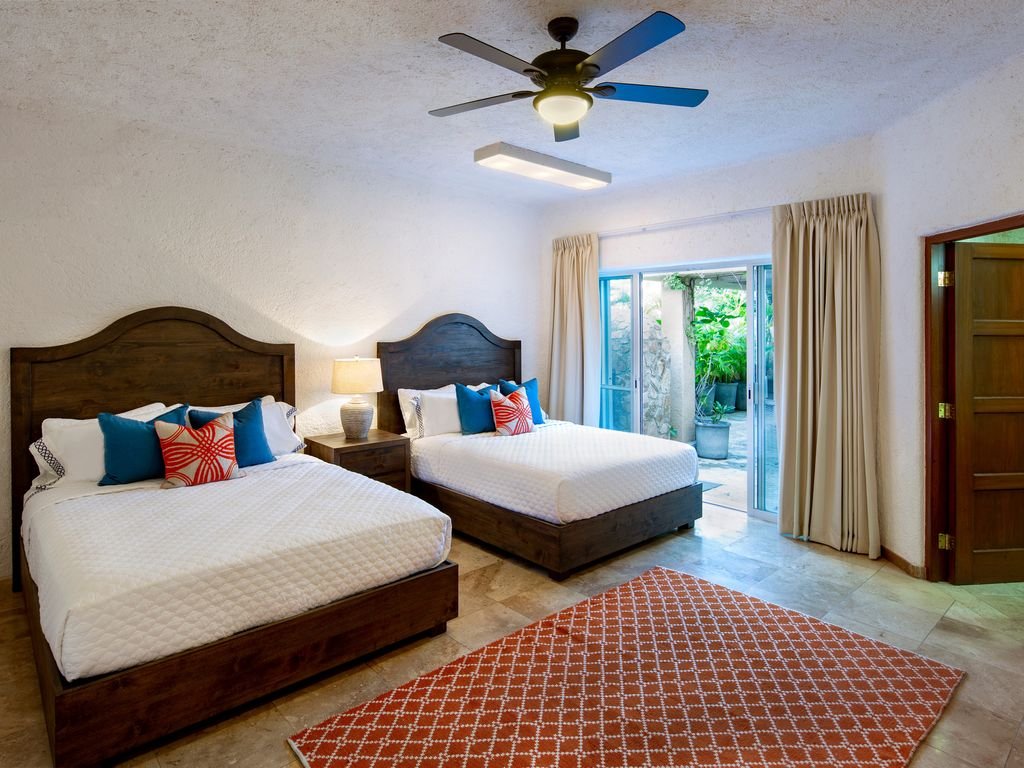 Hotel room with two beds, ceiling fan, red geometric rug, sliding glass doors, and potted plants outside.