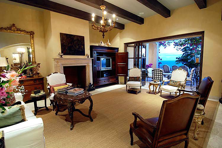 Luxury living room with classic furniture, chandelier, fireplace, and a view of a patio with outdoor seating and ocean scenery in the background.