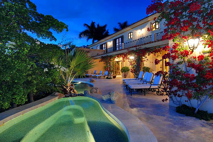 Luxury villa with a pool and outdoor seating area, surrounded by lush greenery and flowering plants, illuminated at dusk.
