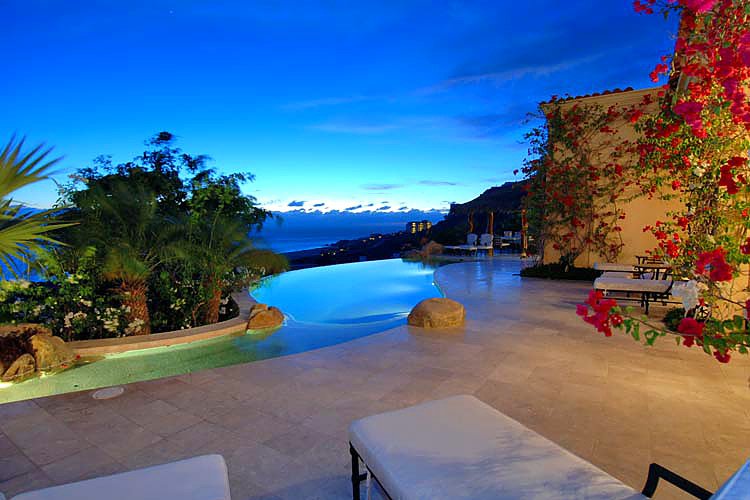 Luxurious patio with infinity pool overlooking ocean and mountains, surrounded by lush greenery and flowers, at sunset.