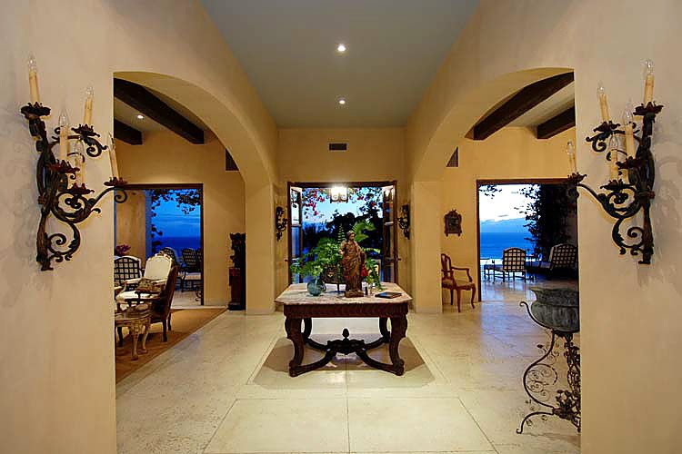 Luxurious open living space with arched doorways, elegant furniture, a central table with decor, candles on walls, and views of an outdoor patio and ocean beyond.