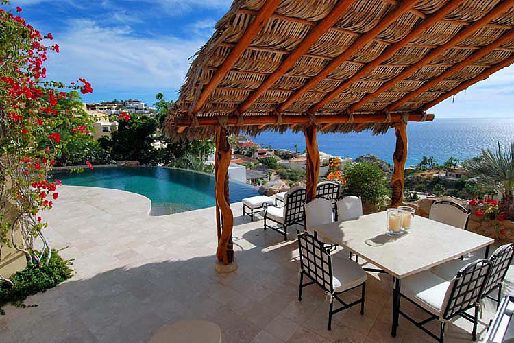 Outdoor patio with a thatched roof, dining table, and ocean view; an infinity pool to the left with bougainvillea vines and scenic landscape.