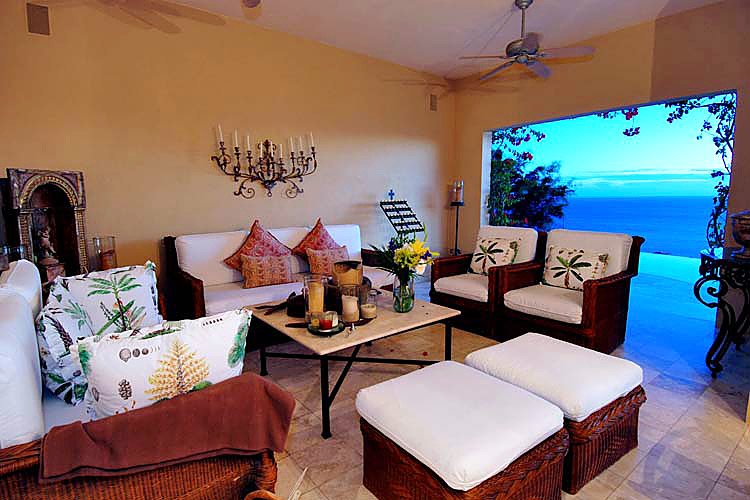 Luxury living room with comfortable wicker furniture, decorative pillows, coffee table with candles and flowers, indoor-outdoor design with ocean view.