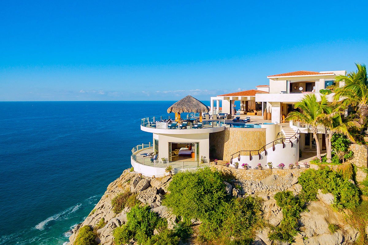 Luxury oceanfront villa on a cliff with a pool and thatched roof structure, overlooking the sea, surrounded by tropical greenery under a clear blue sky.