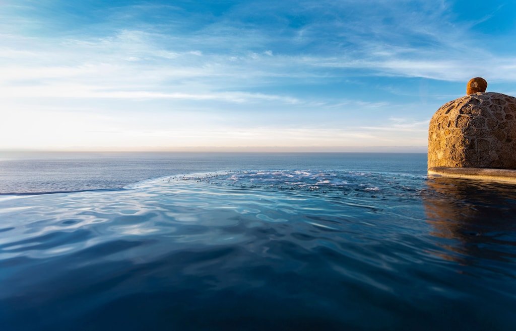 Infinity pool overlooking the ocean with a stone structure and a clear blue sky.