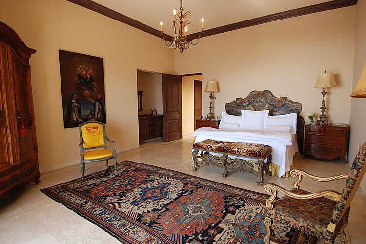 Elegant bedroom with a large bed, ornate headboard, and antique furniture, including a wooden armoire, chairs, and nightstands. A Persian-style rug covers the floor, and a chandelier hangs from the ceiling.
