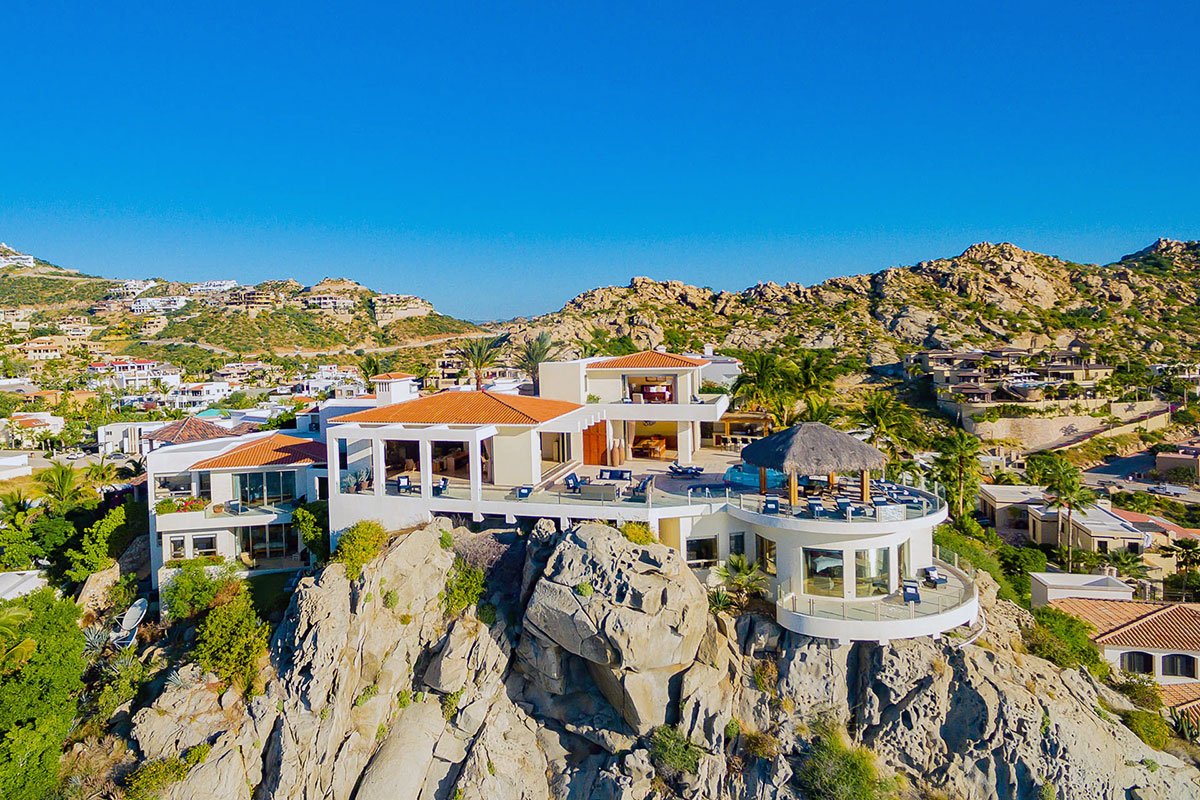 Luxury villa on a rocky hillside with ocean view, surrounded by smaller homes and lush greenery under a clear blue sky.