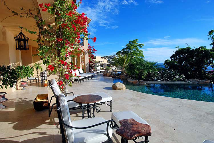 Outdoor patio with lounge chairs, small tables, flowering plants, view of a pool, trees, and distant hills under a clear blue sky.