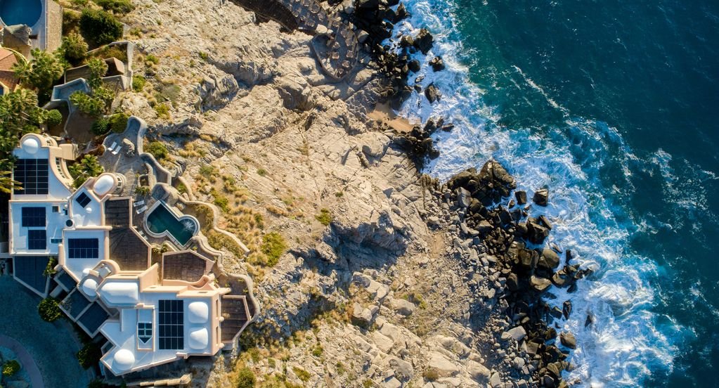 Aerial view of a luxury coastal villa with solar panels, rocky shoreline, and ocean waves.