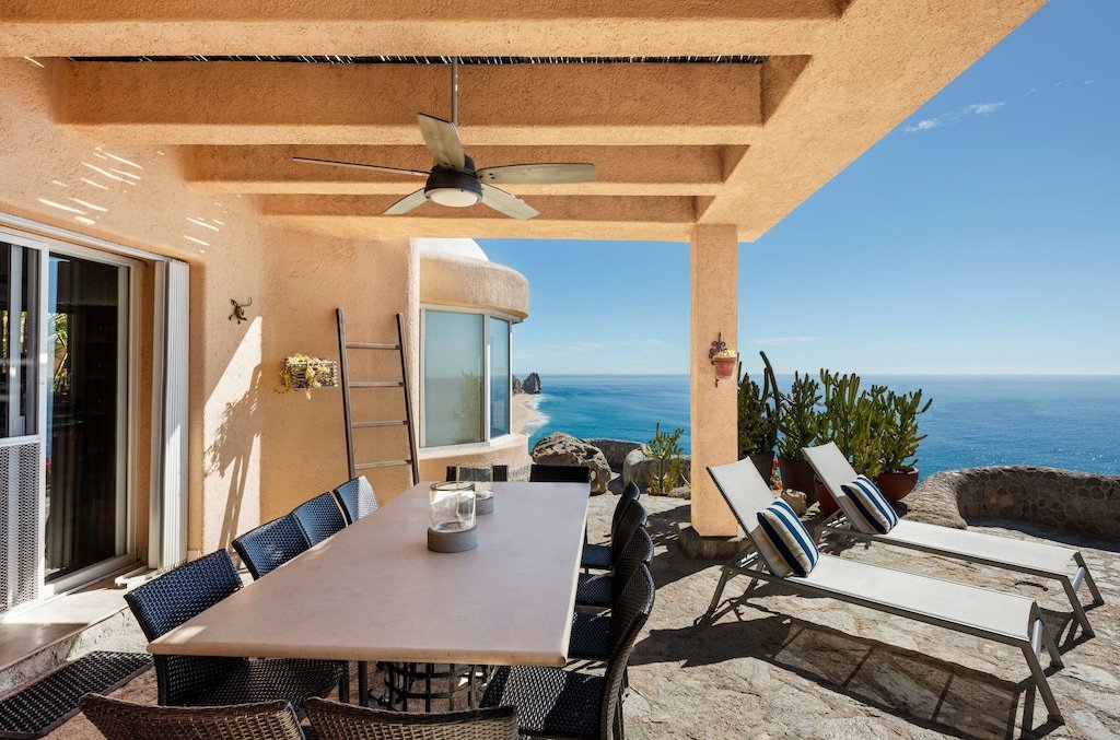 Outdoor patio with a dining table and chairs, two reclining lounge chairs with striped pillows, a ceiling fan, and a view of the ocean. The area is shaded by a pergola, and there's a cactus plant nearby.