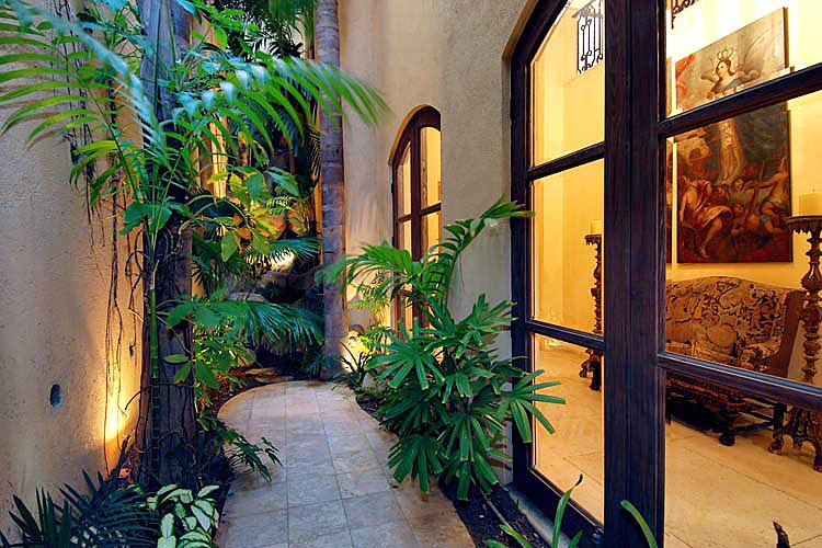 Narrow garden walkway with tropical plants beside a house with large windows and stone flooring.