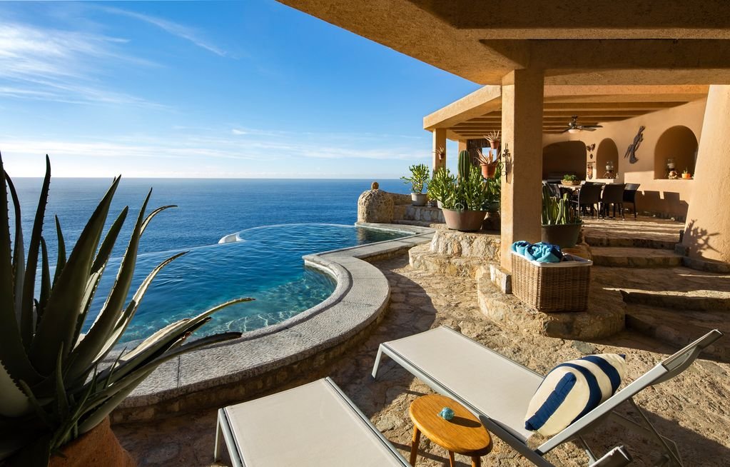 Oceanfront patio with infinity pool, lounge chairs, and covered dining area, overlooking the sea with clear blue sky.