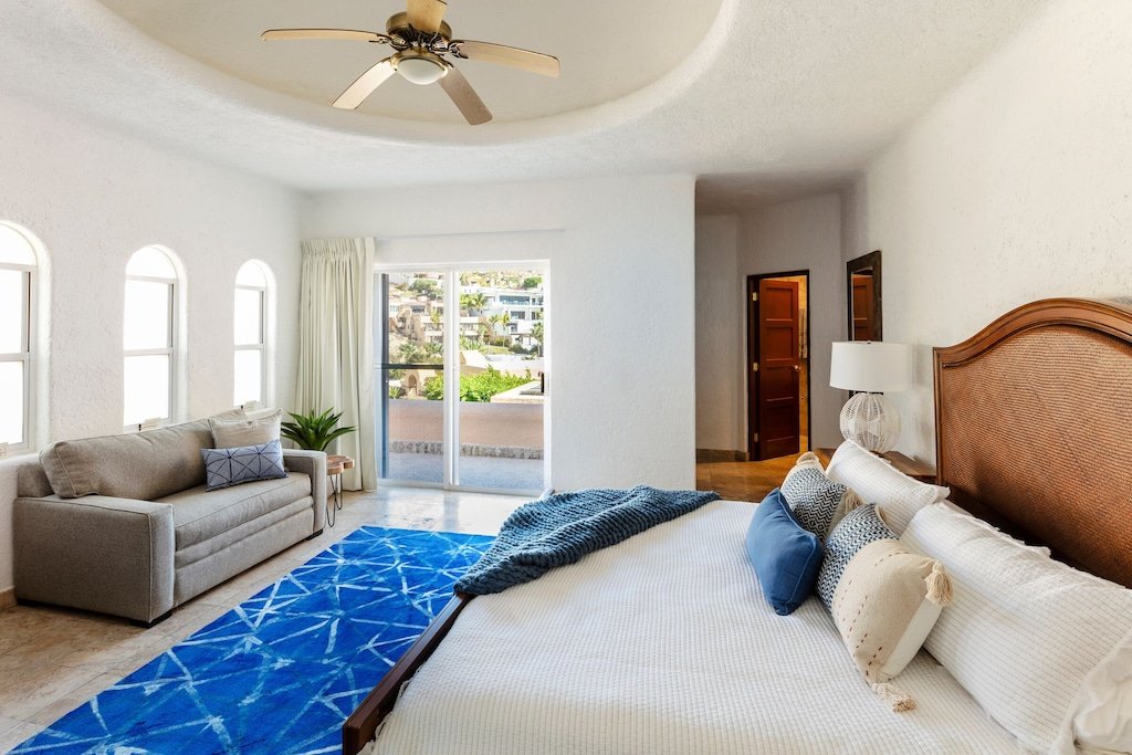 Spacious bedroom with a ceiling fan, large bed, sofa, blue area rug, and a window view, decorated in neutral tones.