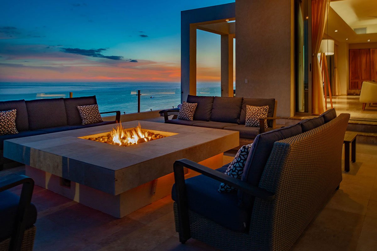 Luxury outdoor patio with fire pit, ocean view at sunset, and modern seating arrangement.