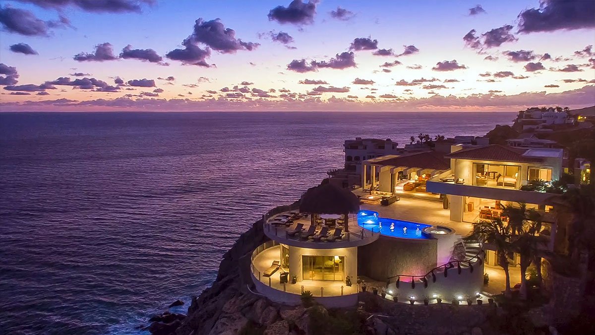 Luxury cliffside resort at sunset overlooking the ocean, featuring illuminated pool and terrace.