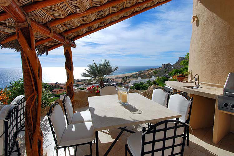 Outdoor dining area with a thatched roof overlooking a coastal view, featuring a marble table, white cushioned chairs, potted plants, and a grill.