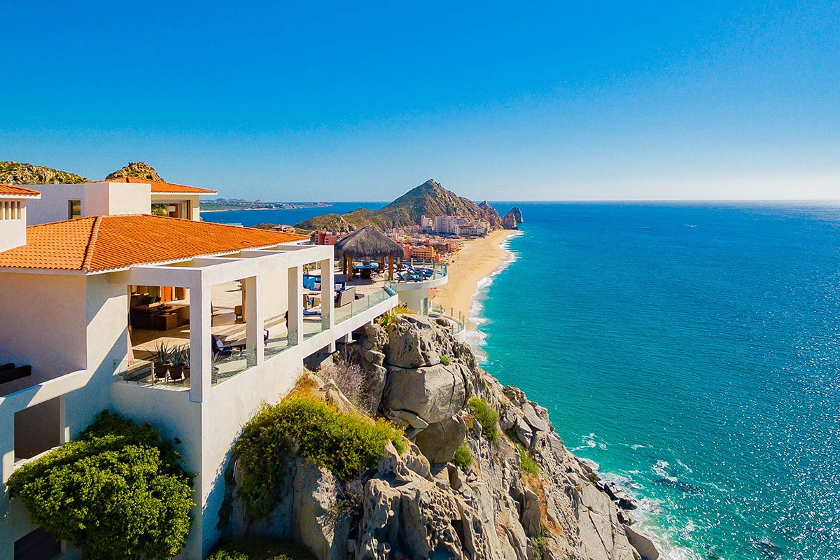 Luxury beachfront villa on a cliff overlooking the ocean, with a sandy beach and distant mountains under a clear blue sky.