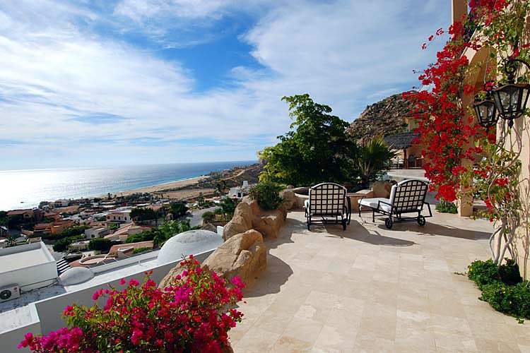 Scenic ocean view from a patio with lounge chairs, vibrant flowers, and a clear blue sky over a coastal town.