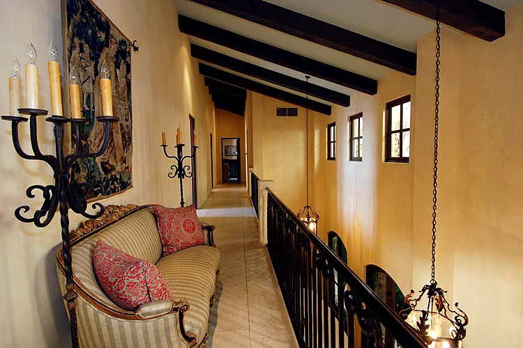 A hallway with exposed wooden ceiling beams, iron light fixtures, a striped couch with red pillows, and wall tapestry.
