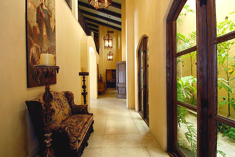 Elegant hallway with beige walls, dark wooden beams, and arched windows. Features ornate furniture, a large painting, and indoor plants visible through the windows.