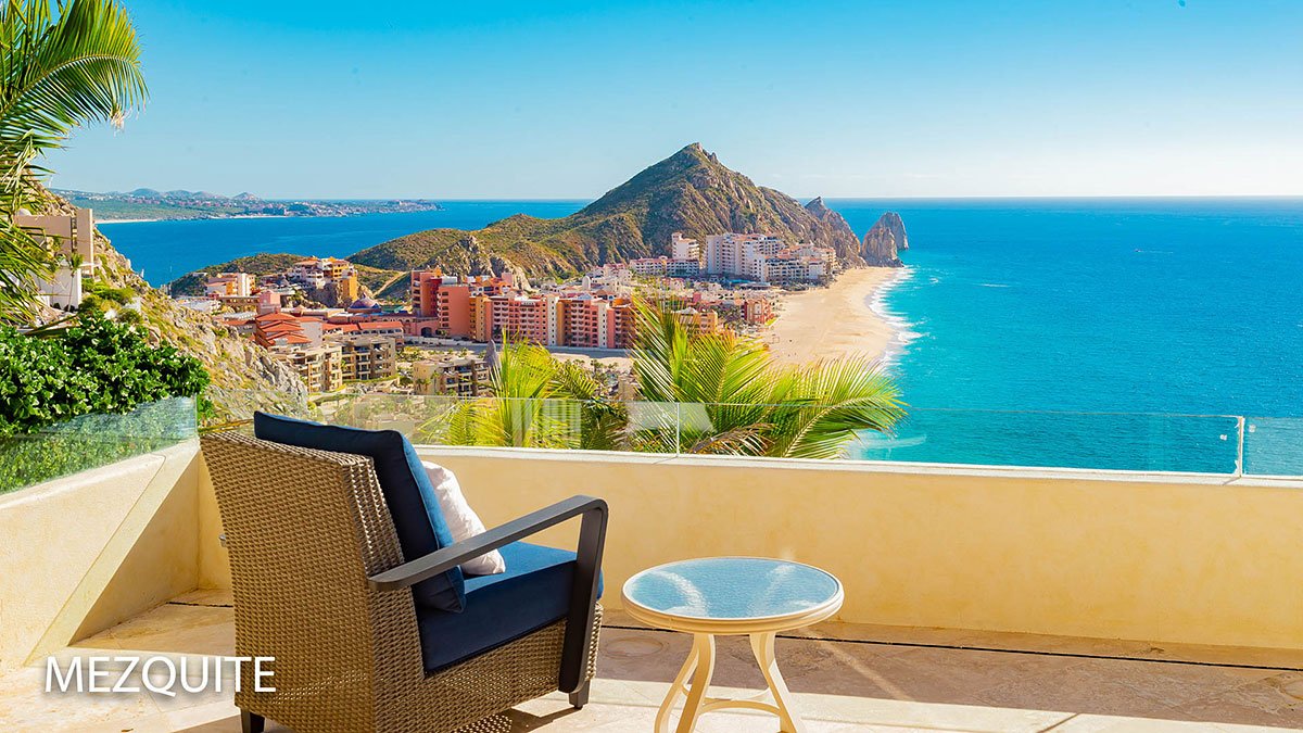 Scenic view from a terrace overlooking a beach with clear blue ocean, buildings, and a rocky hillside in the distance.