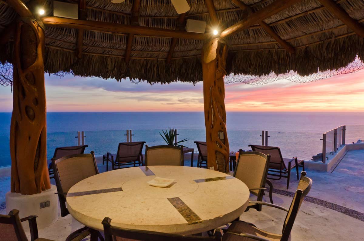 Outdoor patio with a round table and chairs under a thatched roof, overlooking a scenic ocean view at sunset.