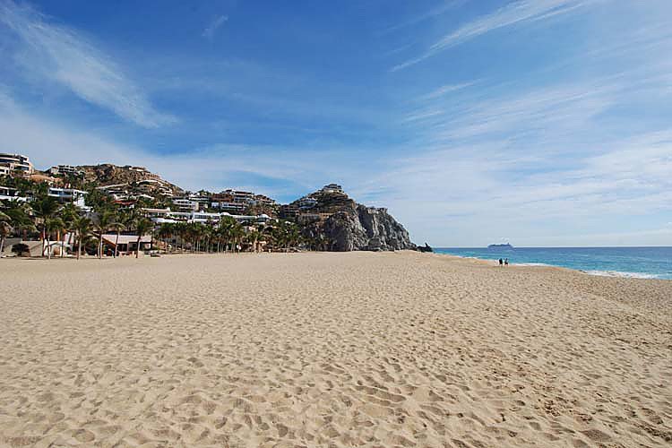 Sandy beach with clear blue sky, rock formation in background, and ocean waves. Distant buildings on hillside.