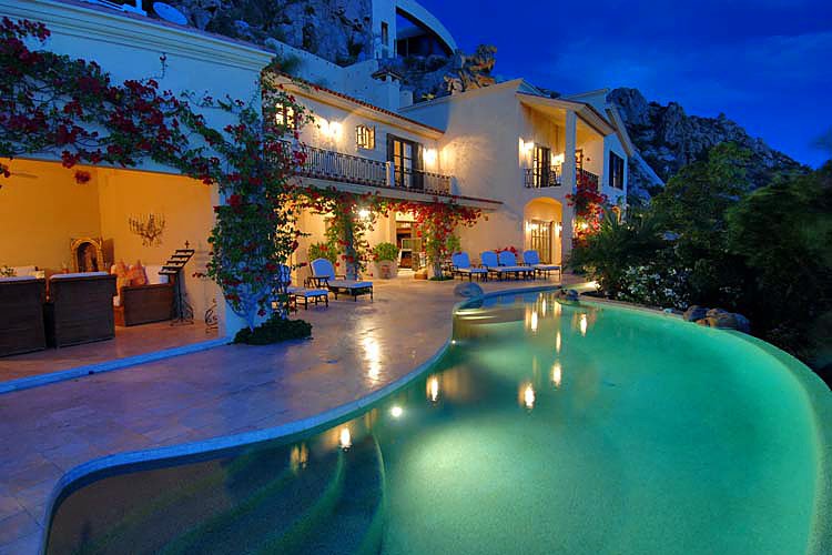 Luxury villa with illuminated pool and patio at dusk, surrounded by lush greenery and mountain views.