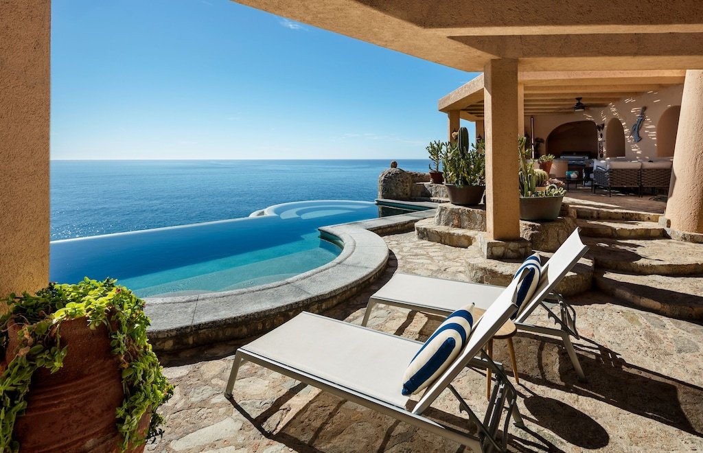 Luxury patio overlooking ocean with infinity pool, lounge chairs, and potted cacti.