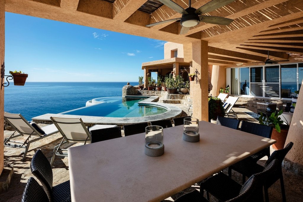 Luxury patio with a view of the ocean featuring an infinity pool, dining table, chairs, and lounge chairs under a pergola.
