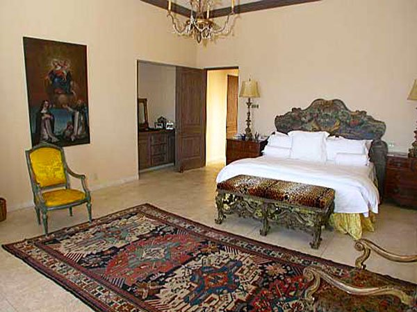 Elegant bedroom with ornate furniture, including a richly patterned area rug, decorative bench, vintage armchair, and a chandelier. Walls are adorned with artwork, and there's a doorway leading to another room.