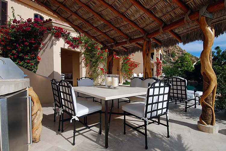 Outdoor patio dining area with thatched roof, white cushioned chairs, and a stone table. Surrounded by blooming flowers and greenery.