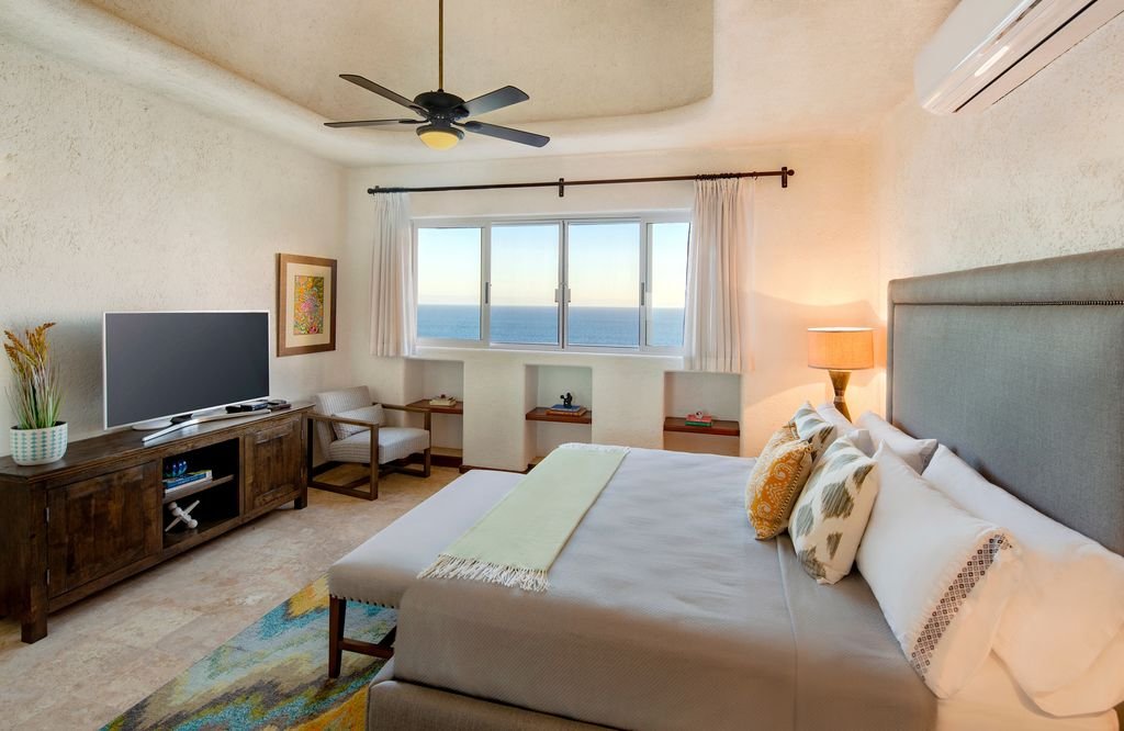 Modern bedroom with ocean view, featuring a bed with decorative pillows, a wooden dresser with a flat-screen TV, a cushioned chair, and a ceiling fan.