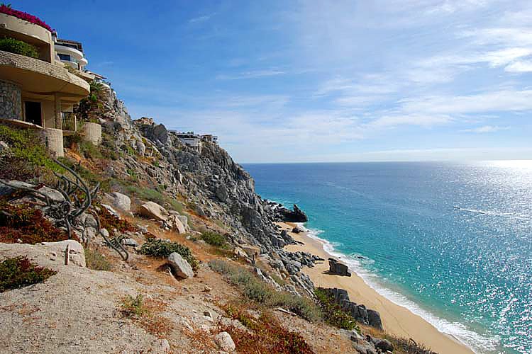 Rocky cliffside with luxury homes overlooking a sandy beach and blue ocean under a partly cloudy sky.