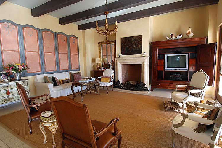 Traditional living room with wooden beam ceiling, beige walls, and a large fireplace. Elegant furniture includes armchairs, a sofa, wooden tables, and a large wooden cabinet with a TV. Decor features a chandelier, framed art, and floral arrangements.