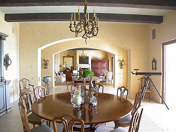 Elegant dining room with a round wooden table, ornate chandelier, and telescope. Arched doorway leading to living area with decorative mirrors and plants.