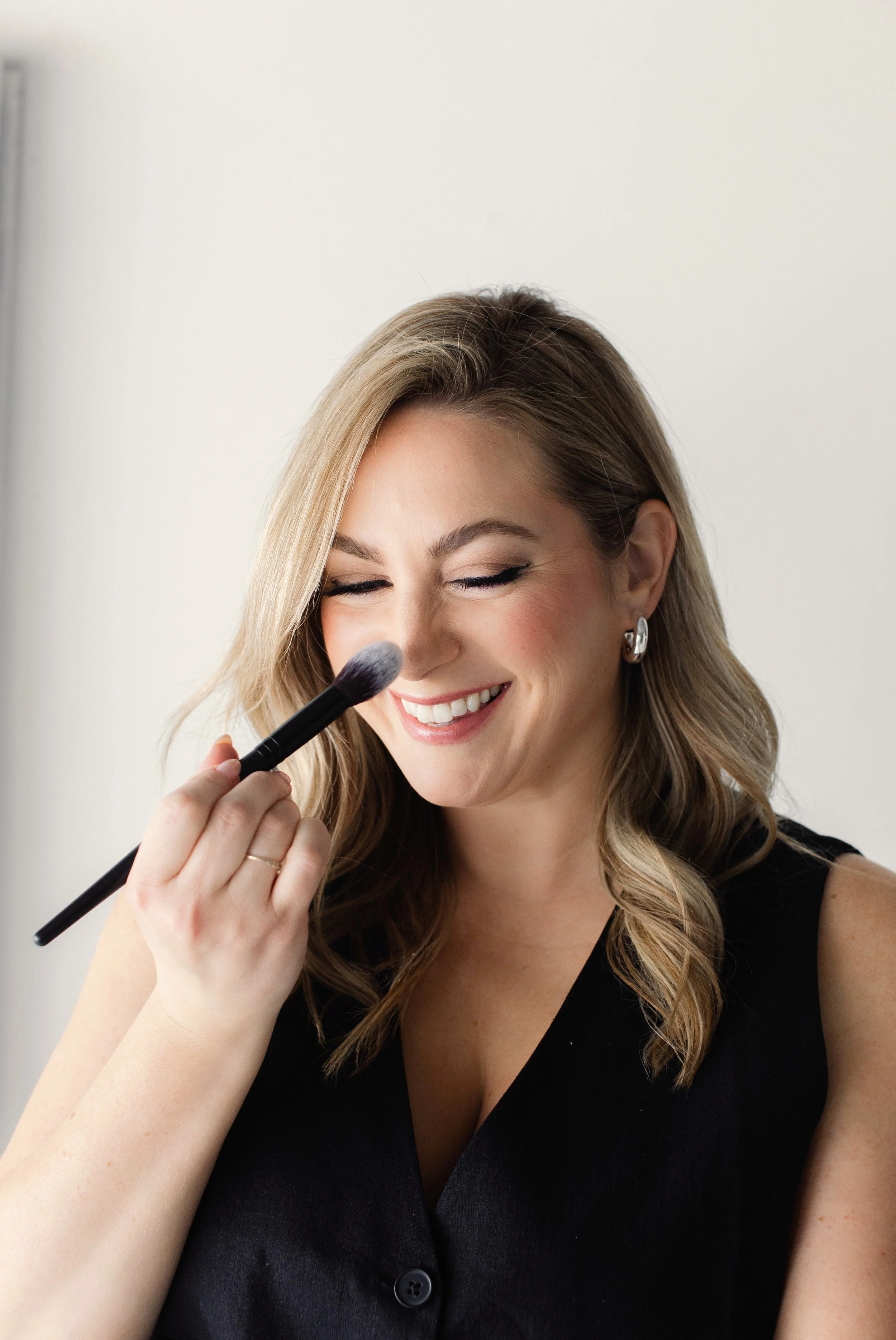 Person smiling while applying makeup with a brush, wearing a black sleeveless top and hoop earrings.