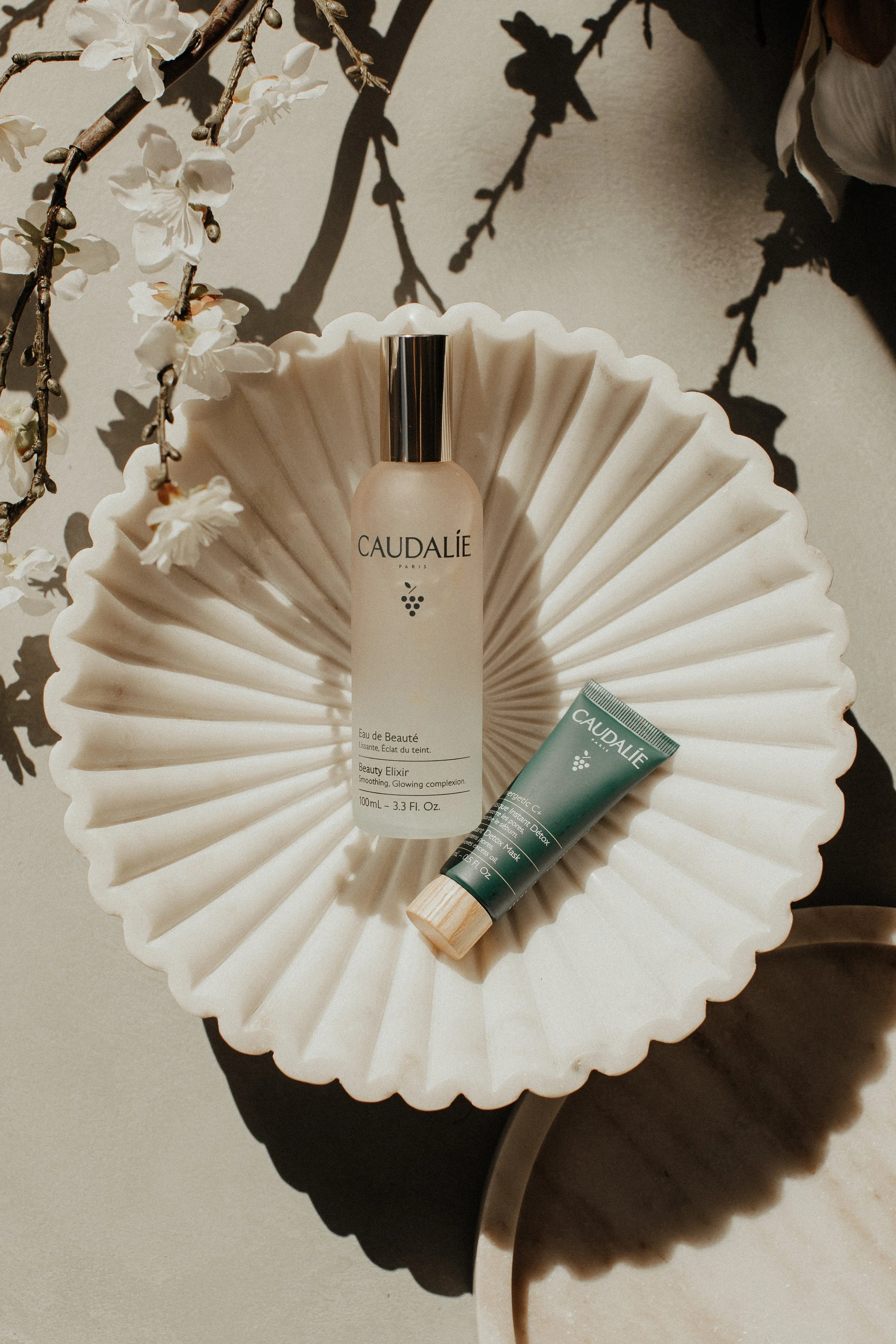 Caudalie skincare products displayed on a fluted ceramic dish with a branch of white flowers as decoration.