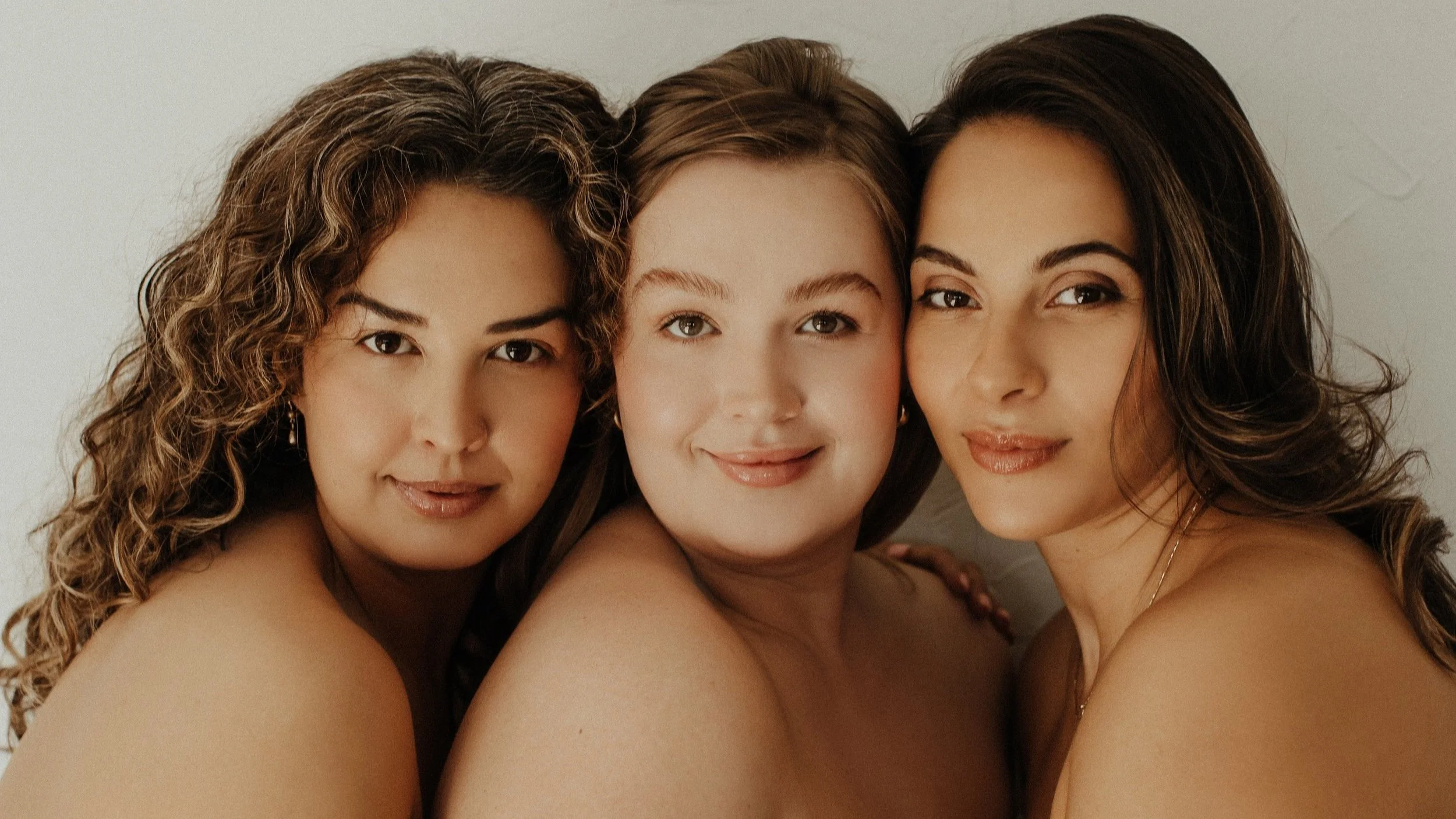 Three women with different hairstyles posing closely, smiling at the camera against a light background.