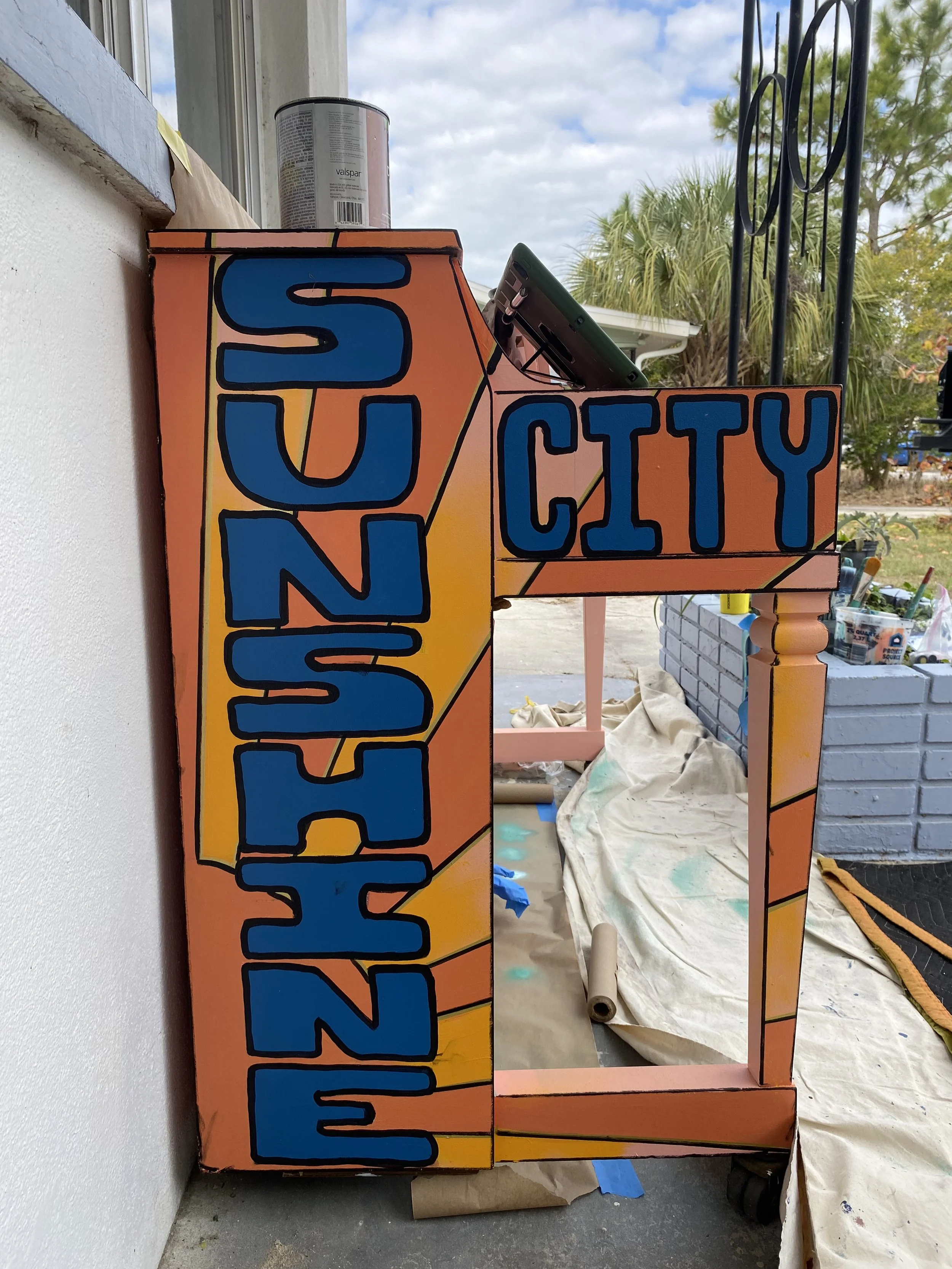 A colorful, hand-painted sign that reads "NASHVILLE CITY" in bold blue letters on an orange background, with some yellow accents and black outlines, leaning against a white wall outdoors.