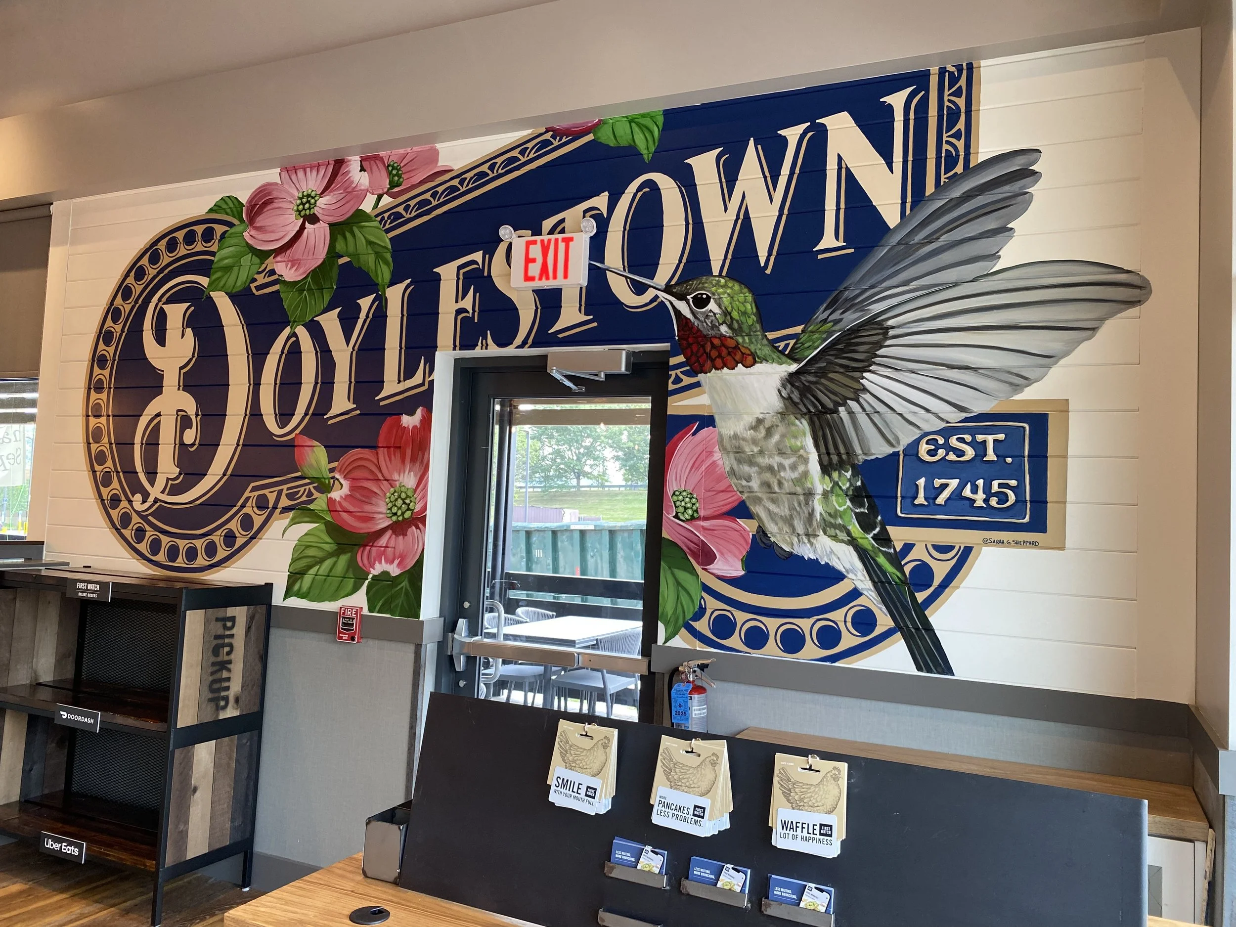A mural on a wall depicting a hummingbird in flight near pink flowers, with the text 'Doylestown' and an 'EST. 1745' in a First Watch Restaurant.