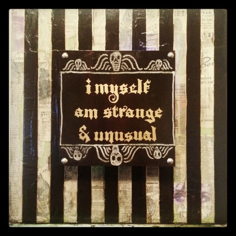 Decorative sign with gothic font saying "I myself am strange & unusual" against a background of vertical black and white striped wallpaper with newspaper print.