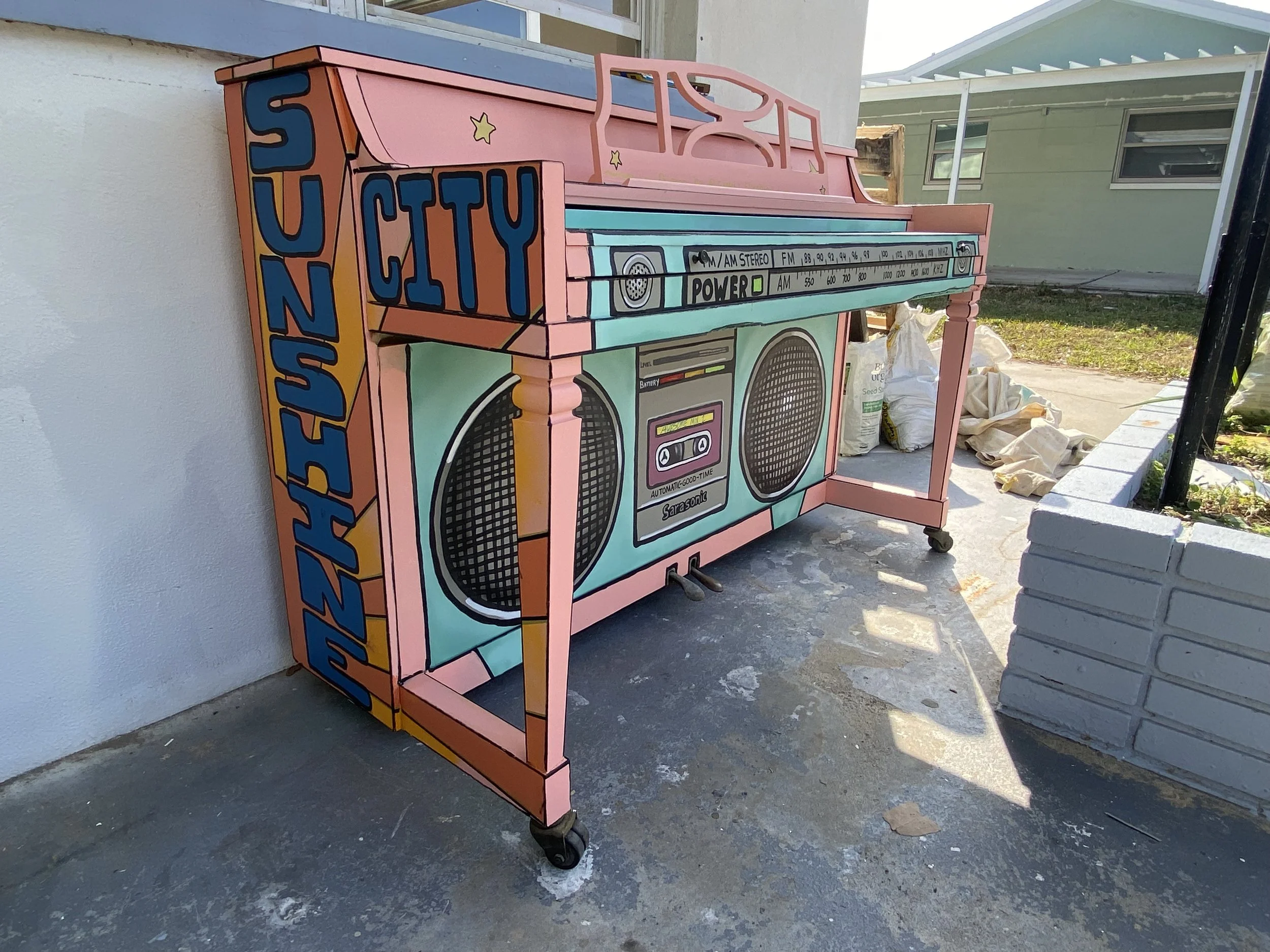 Colorful Piano painted like a 1980s boombox with painted  speakers, and text, placed outdoors on concrete ground near a white wall and garden area.