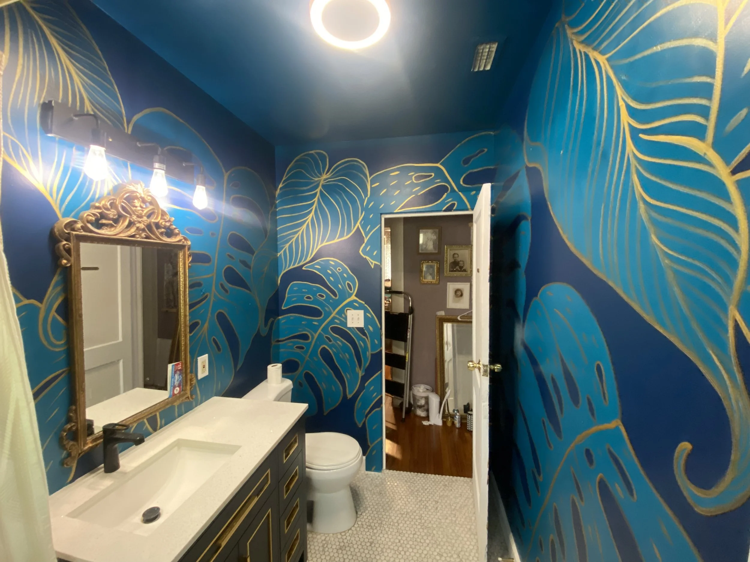 Bathroom with blue and gold leafy mural, gold-framed mirror, white vanity with black fixtures, toilet, and view into a hallway with framed pictures.