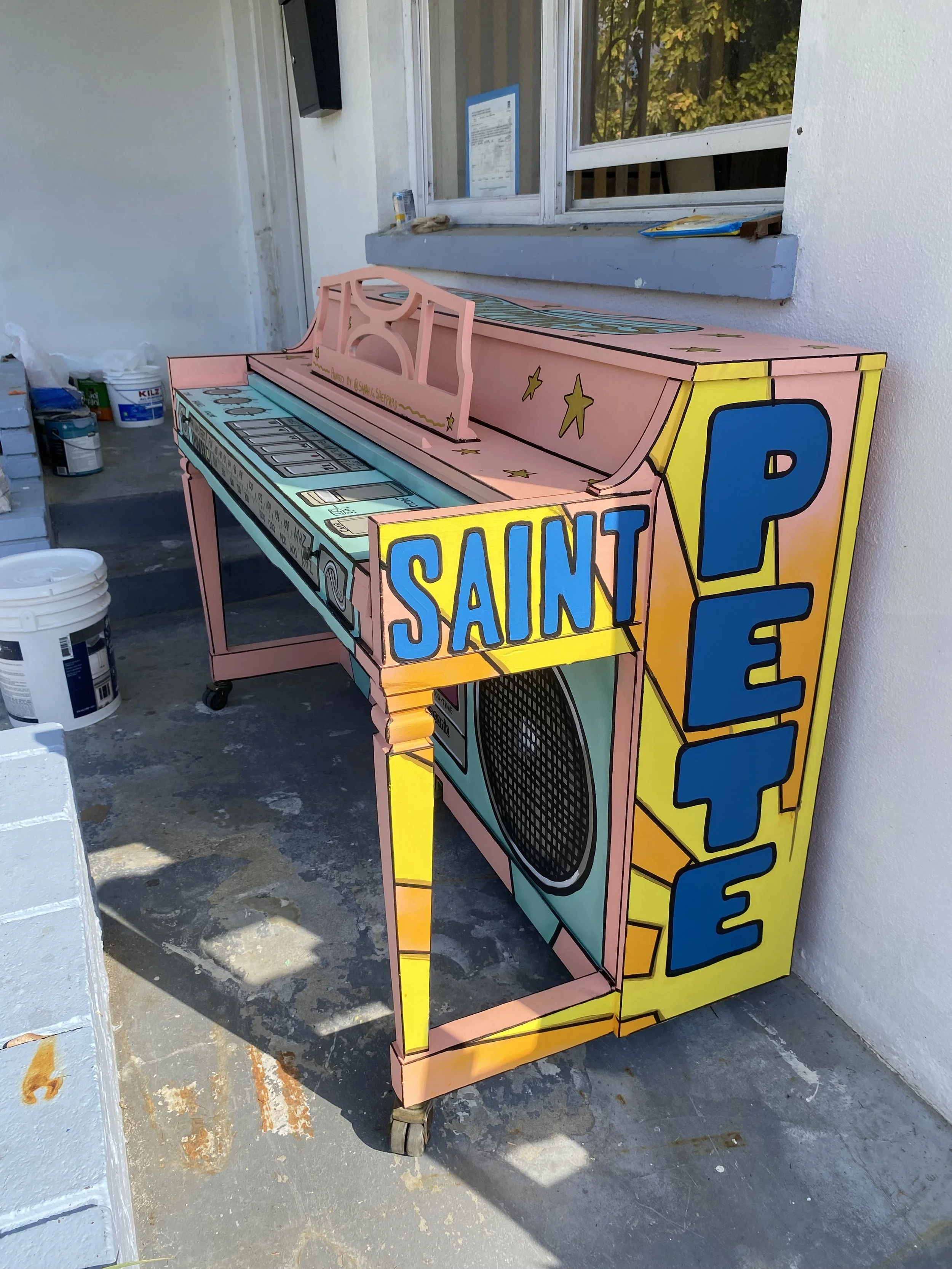 Colorful painted piano game machine with the words 'Saint Pete' on side, positioned outside next to a concrete wall and stairs.