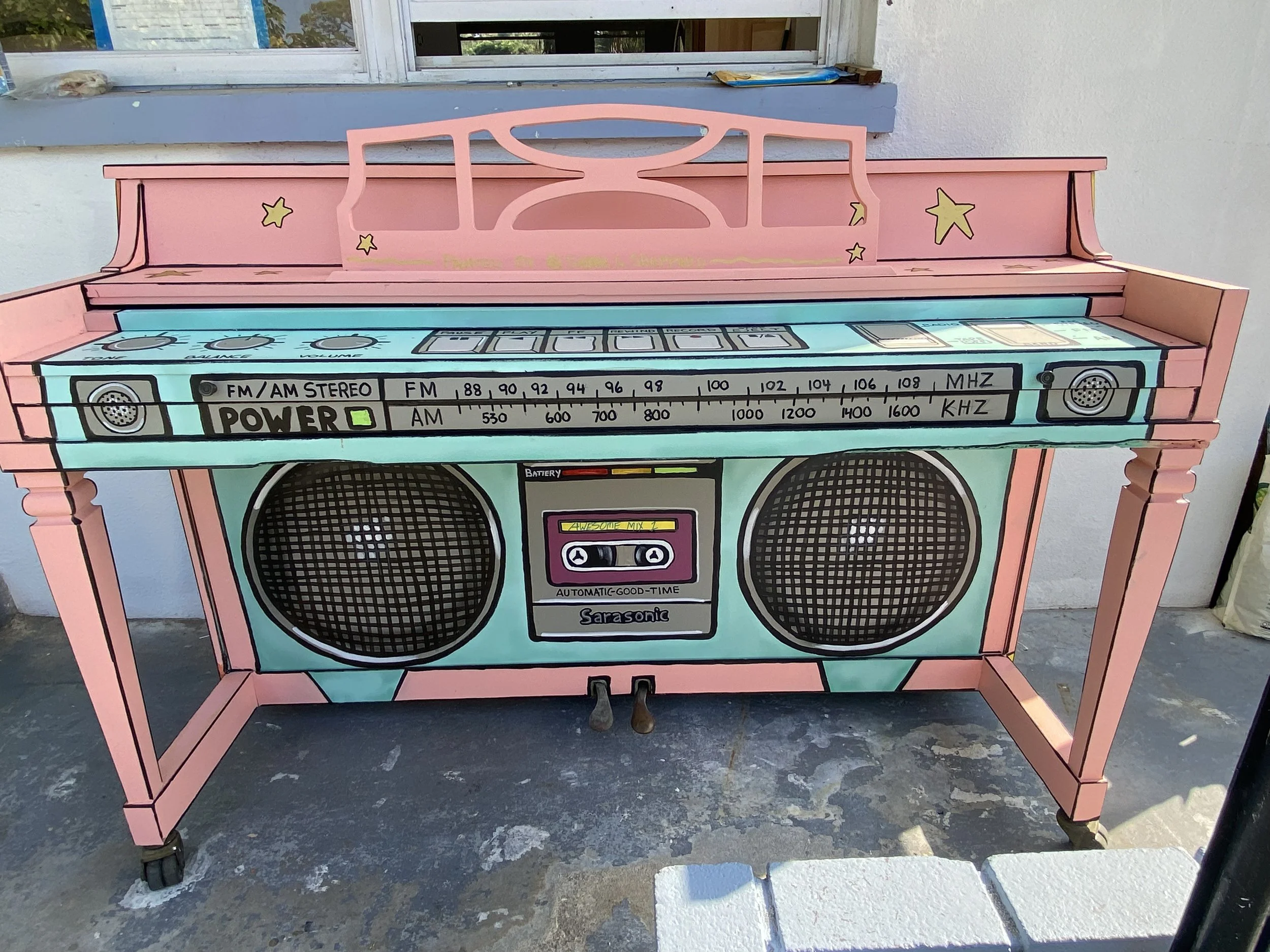 Pink painted cardboard play stand shaped like a vintage boombox with colorful details and stars, designed for children, placed on a concrete surface outside.