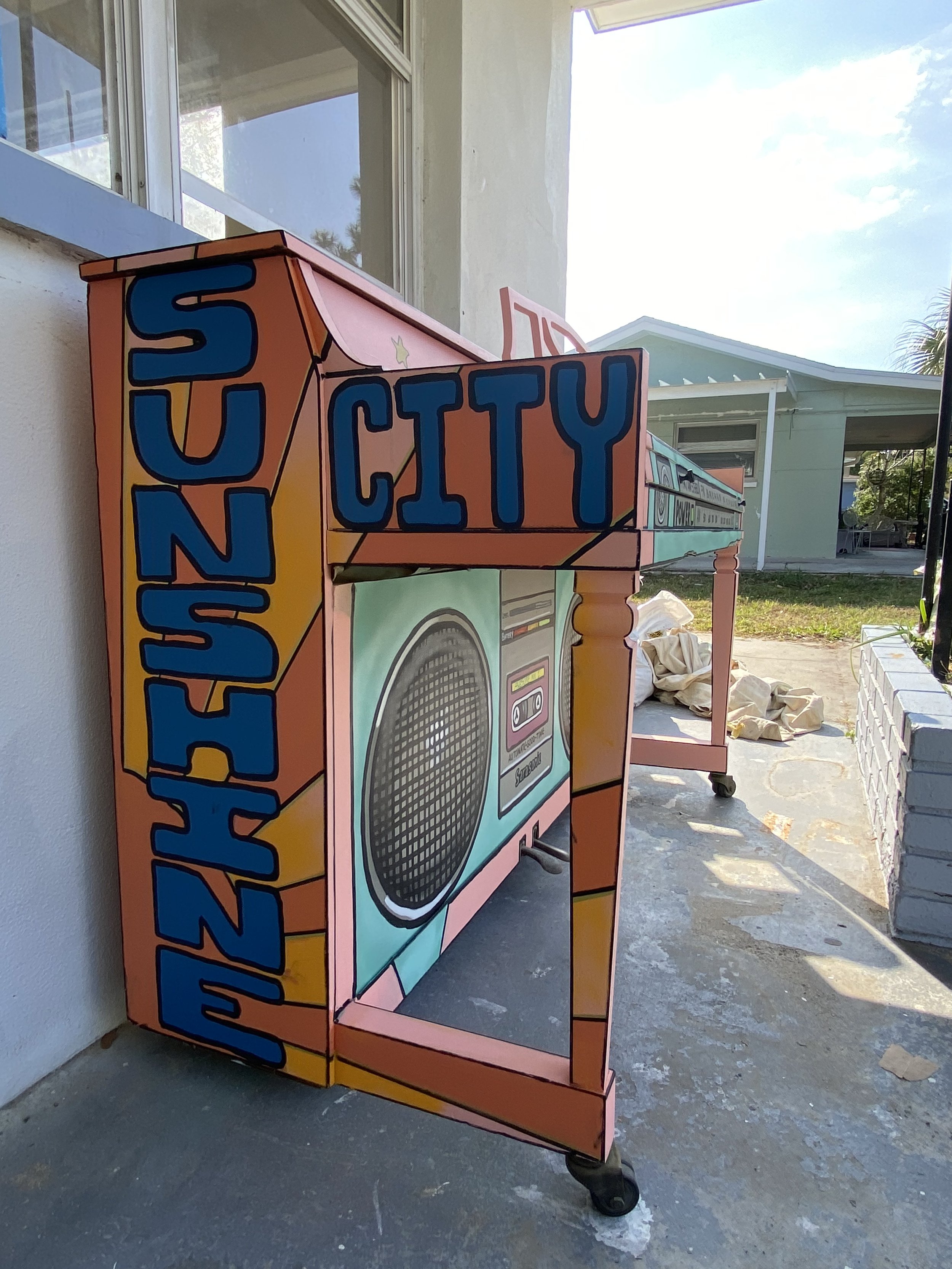 Colorful cardboard box decorated with a retro theme, featuring a large speaker and washing machine illustration, with words "Suntory" and "City" on the sides and top, on a porch outside a house.